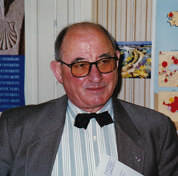 Portrait d’Yvan Tchéfranoff, ancien Secrétaire Général de l’Université de Limoges. Il porte des lunettes et un nœud papillon noir, vêtu d’une chemise rayée et d’une veste grise, posé devant une carte et des documents accrochés au mur.