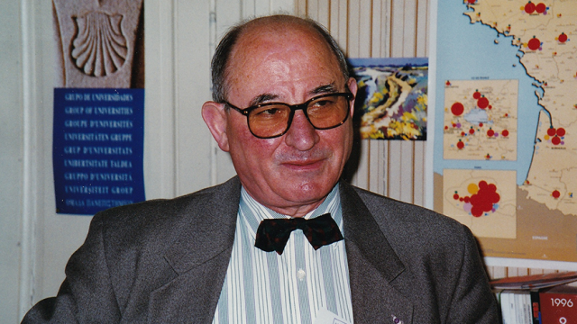 Portrait d’Yvan Tchéfranoff, ancien Secrétaire Général de l’Université de Limoges. Il porte des lunettes et un nœud papillon noir, vêtu d’une chemise rayée et d’une veste grise, posé devant une carte et des documents accrochés au mur.