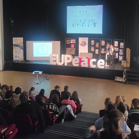 Vue d’ensemble d’une conférence du festival EUPeace à l’Université de Limoges. Le public est assis dans un amphithéâtre, face à une scène où un intervenant présente un diaporama. De grandes lettres formant le mot « EUPeace » sont installées sur la scène, encadrées par deux statues de vaches rouges, symboles de la région limousine.