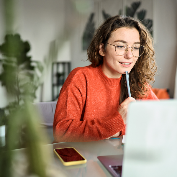 Jeune femme portant un pull orange travaillant sur un ordinateur portable depuis chez elle, concentrée et souriante.