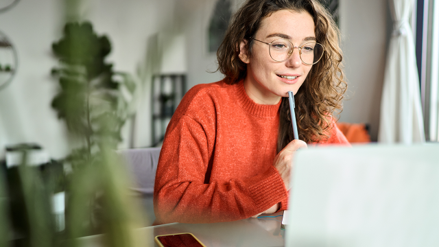 Jeune femme portant un pull orange travaillant sur un ordinateur portable depuis chez elle, concentrée et souriante.