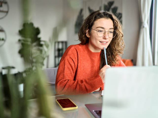 Jeune femme portant un pull orange travaillant sur un ordinateur portable depuis chez elle, concentrée et souriante.