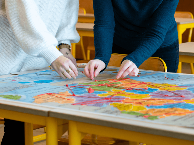 Deux personnes debout autour d’une carte du monde en salle de classe, reliant différents pays avec du fil rouge pour illustrer des échanges internationaux.